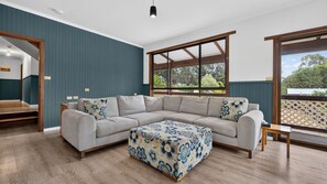 A modern living room with a sectional sofa, a patterned ottoman, and a large window offering a view of greenery. - Treetop Retreat House | Spacious Family Getaway (Metung)