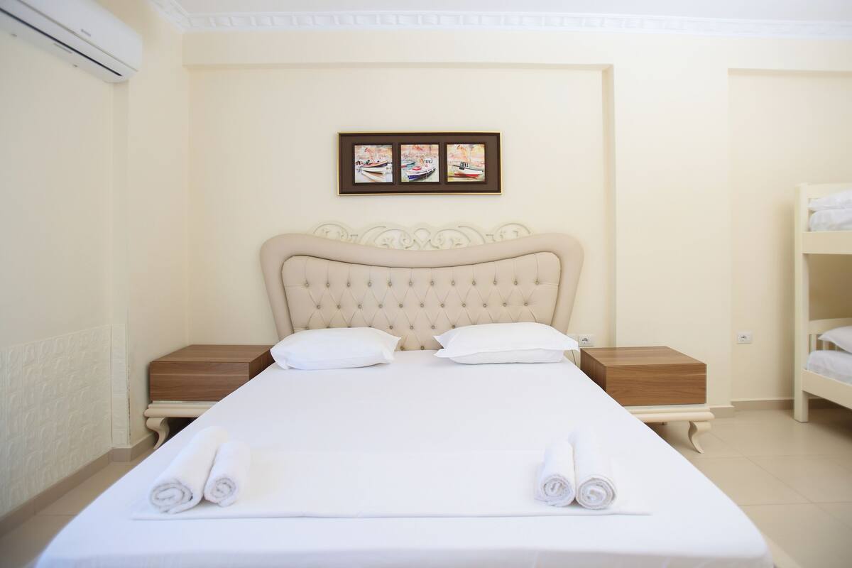 A neatly made bed with white linens and pillows, a wooden nightstand, a framed picture on the wall, and a bunk bed to the right.