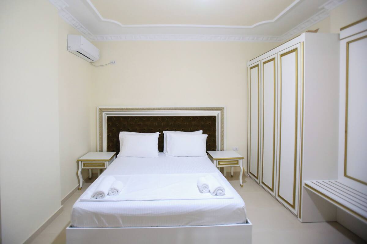 A neatly made bed with white linens and two rolled towels, a wooden headboard, and a white nightstand with a lamp.