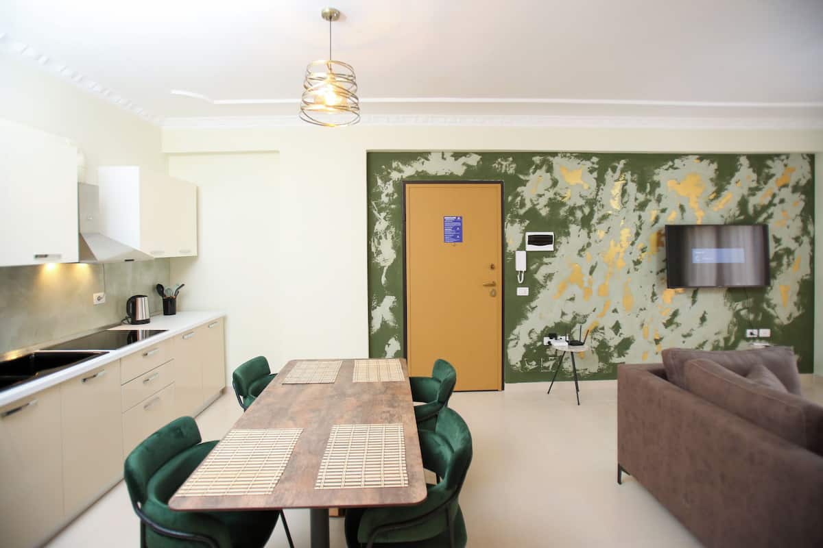 A compact living space with a kitchenette, a dining area with a wooden table and green chairs, a sofa, and a television mounted on the wall.