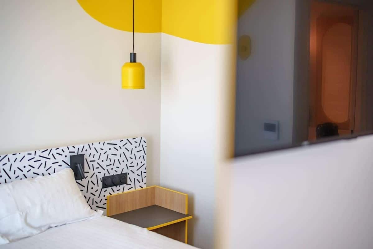 Une chambre moderne avec un lit blanc, une tête de lit à motifs, une table de chevet en bois et une lampe suspendue jaune.