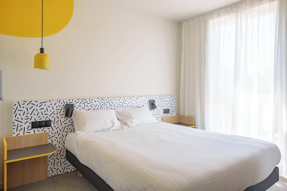 Une chambre d’hôtel moderne avec un grand lit, une table de chevet, une petite table et une lampe suspendue jaune.