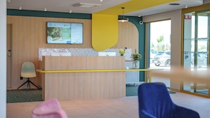 Un espace d’accueil avec un comptoir de réception en bois, une chaise bleue et un mur d’accent jaune.