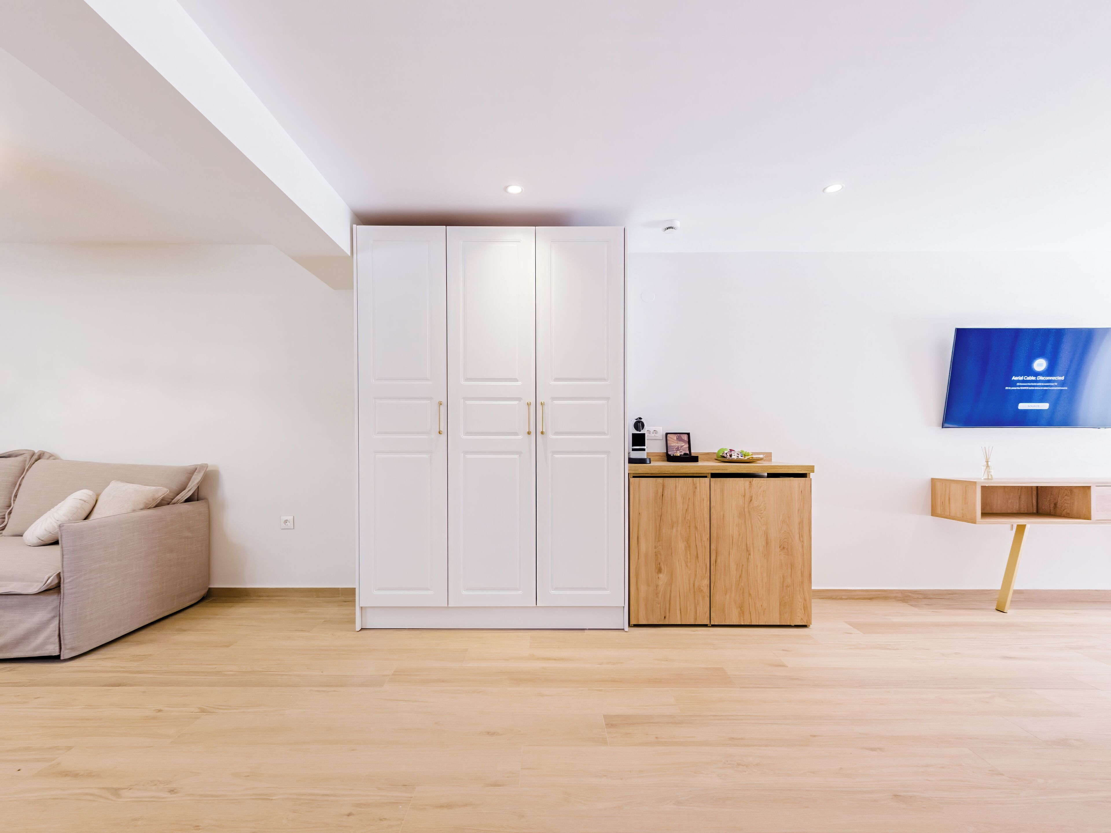 Una sala de estar moderna con un sofá color beige, un armario blanco, un mueble de madera y un televisor montado.