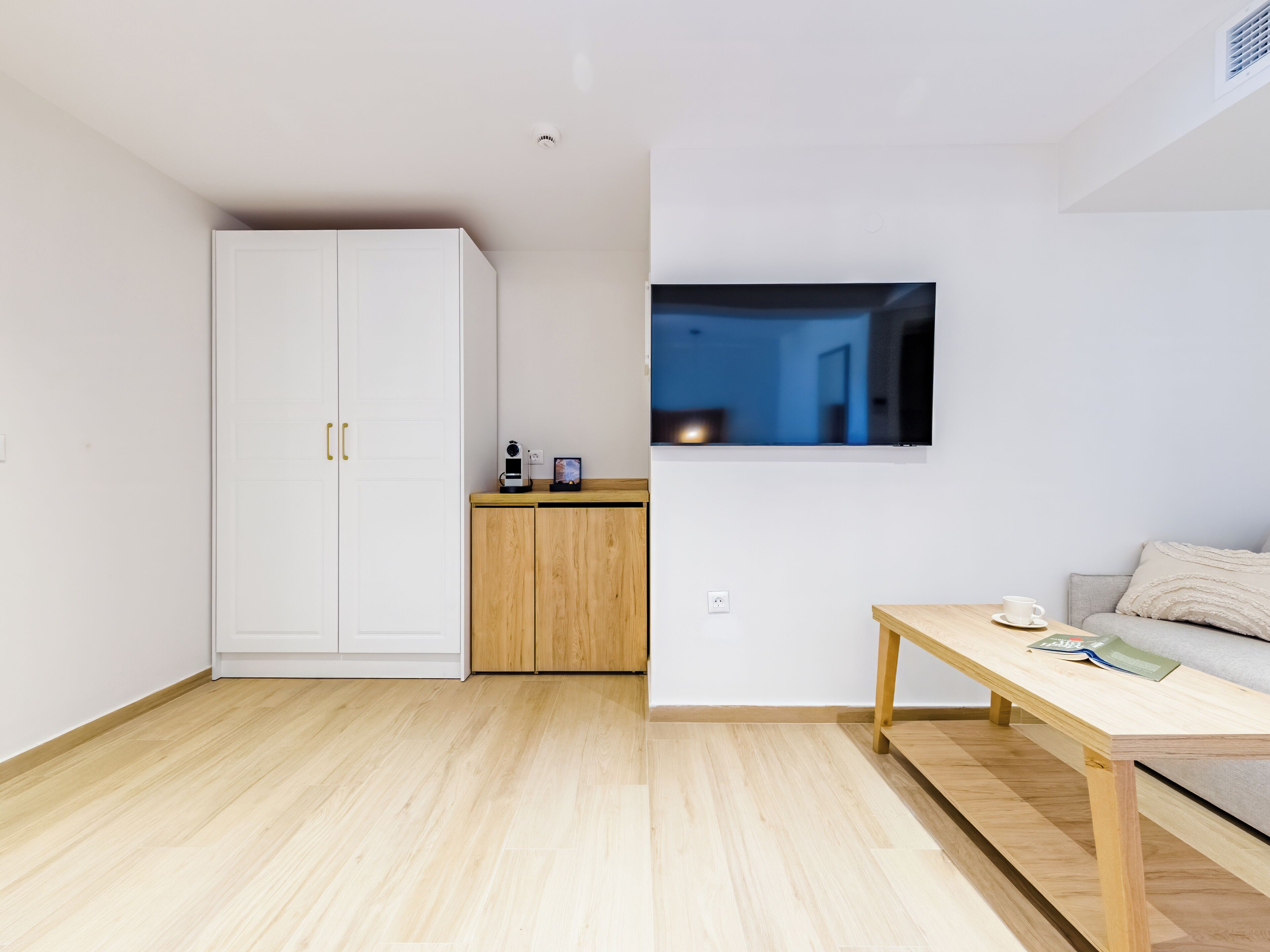 Une chambre moderne et minimaliste, avec une armoire blanche, un meuble en bois, un téléviseur fixé au mur et une table en bois sur laquelle se trouvent une tasse et un magazine.