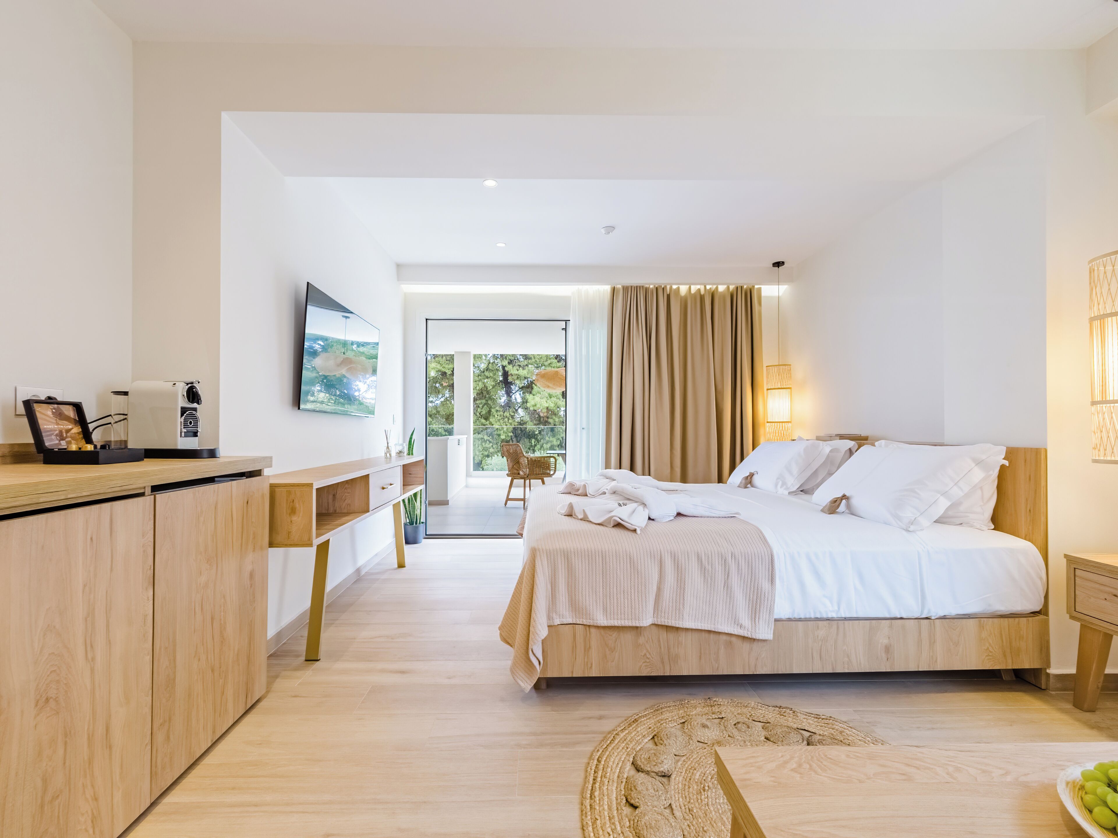 A modern hotel room with a large bed, a desk with a coffee machine, a TV, and a view of the outdoors.
