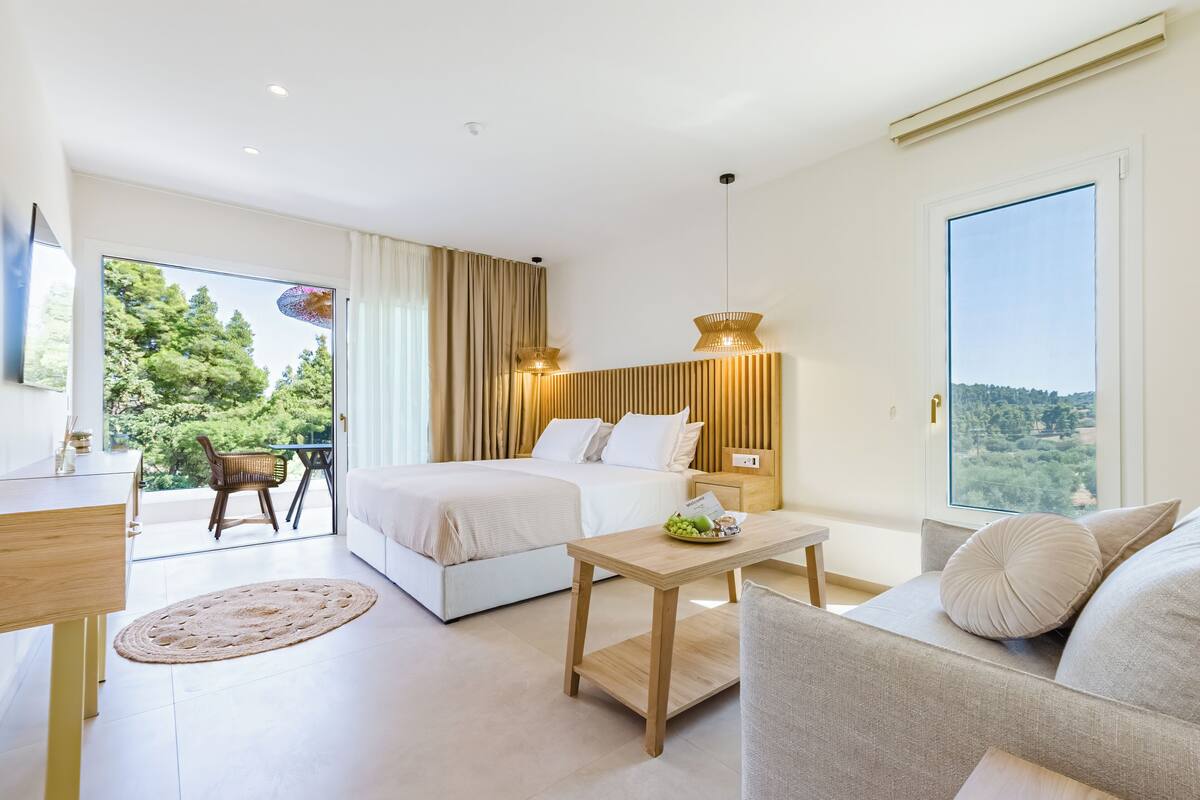 A modern hotel room with a bed, a sofa, a small table, and a balcony with a view of the trees.