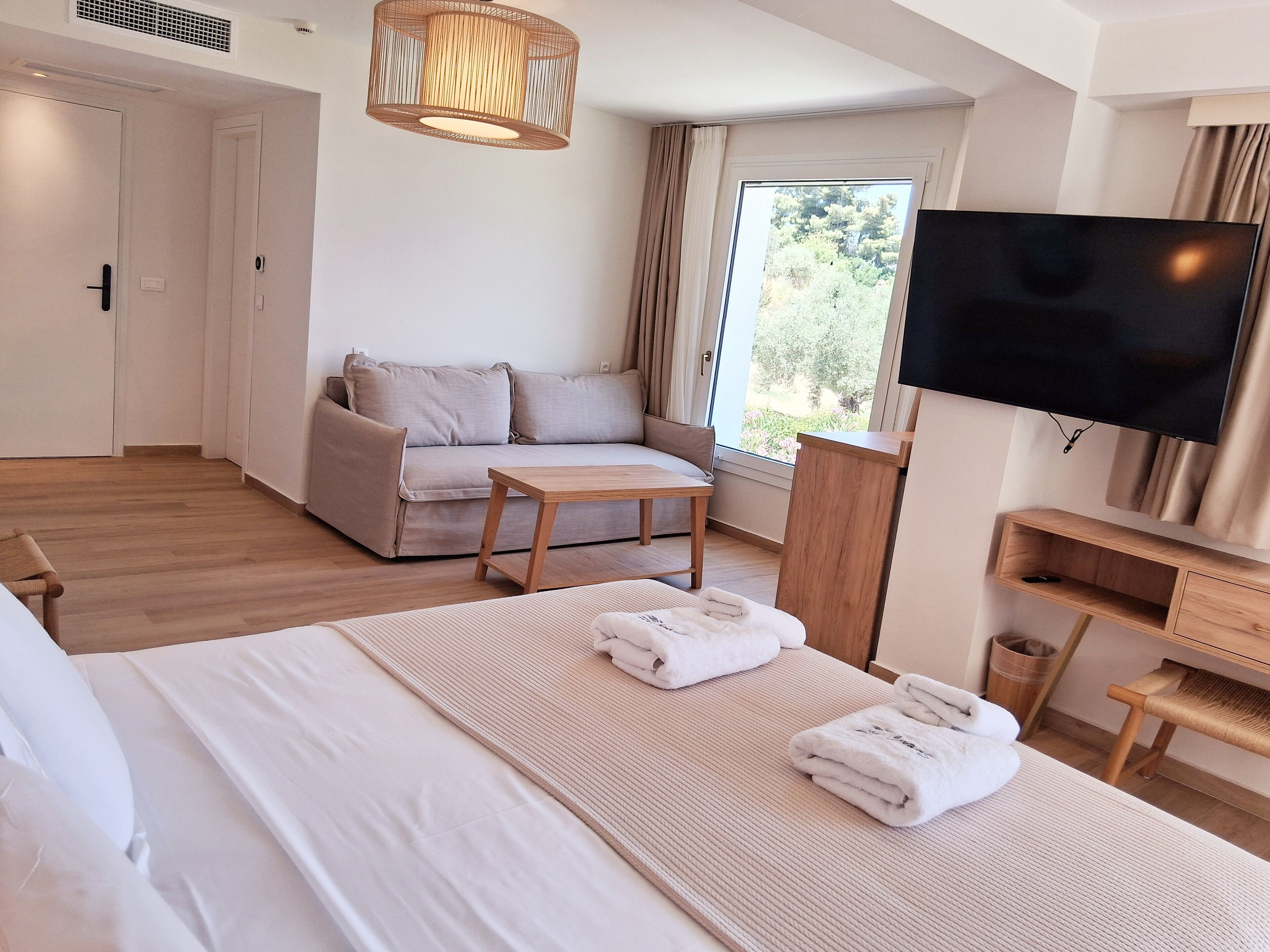 A modern hotel room with a bed, a sofa, a small table, a TV, and a window with a view of trees.