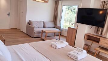A modern hotel room with a bed, a sofa, a small table, a TV, and a window with a view of trees.