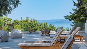 Terraza con sillones de sol y una hamaca, con vistas a un cuerpo de agua y vegetación.