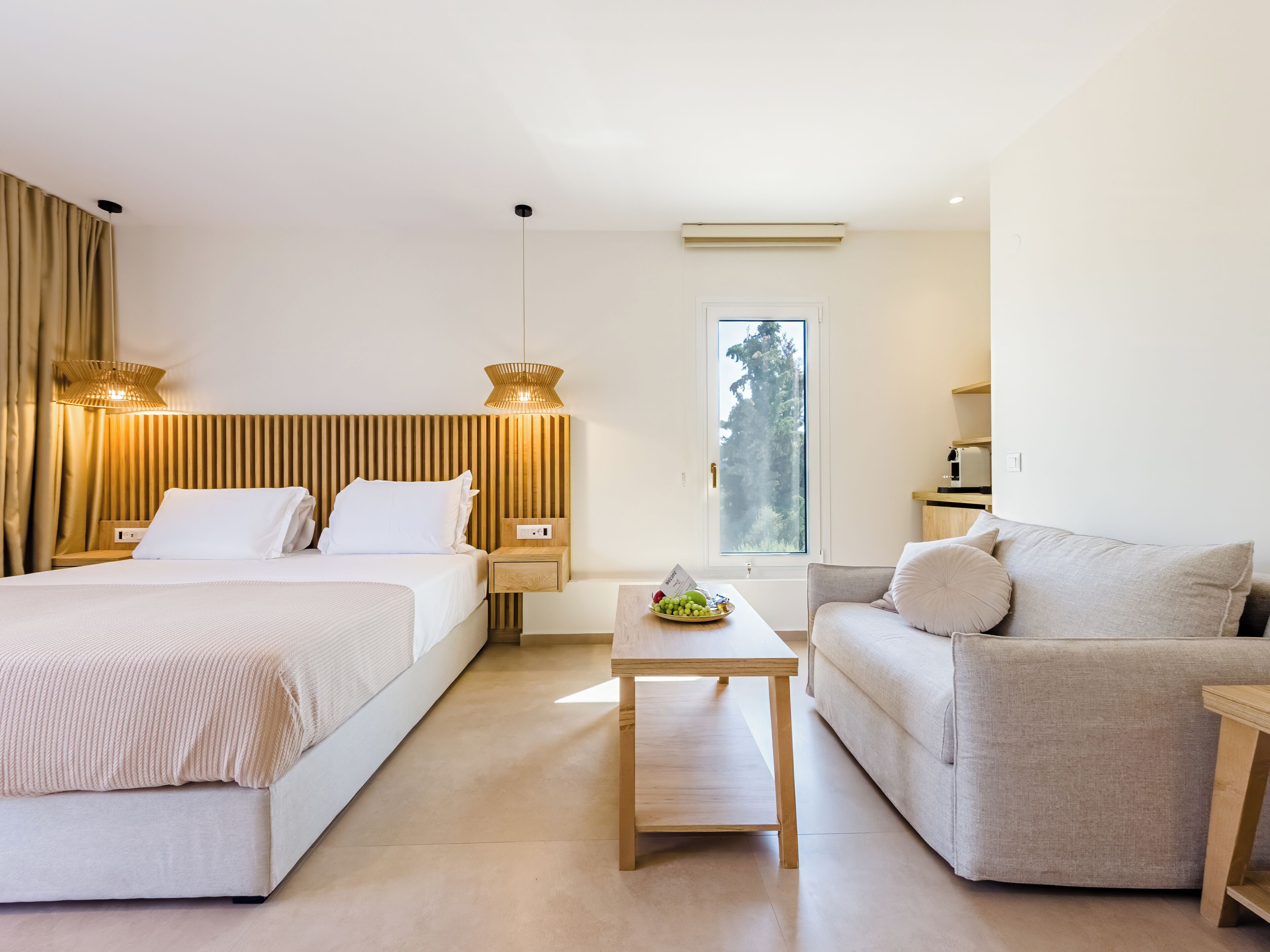A hotel room with two beds, a sofa, a small table, and a window with a view of trees.