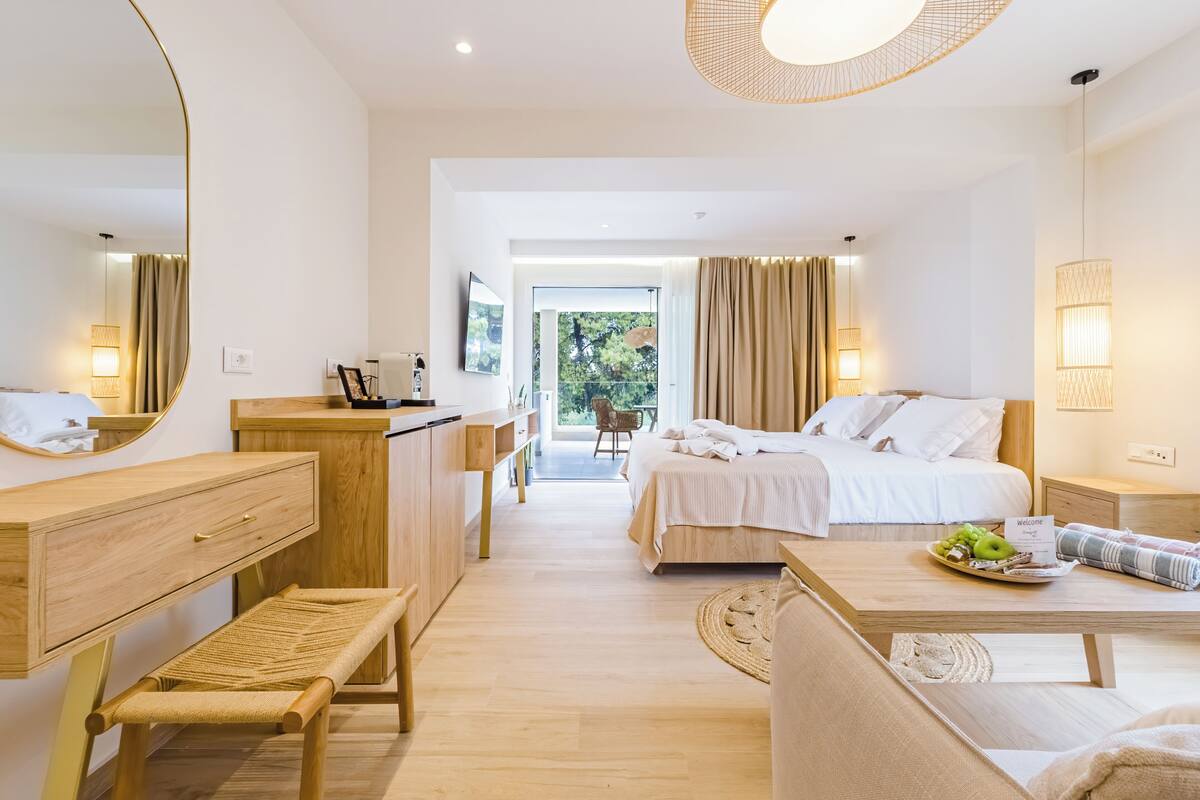 A modern hotel room with a wooden desk, a bed with white linens, a bench, and a dining table with a plate of fruit.