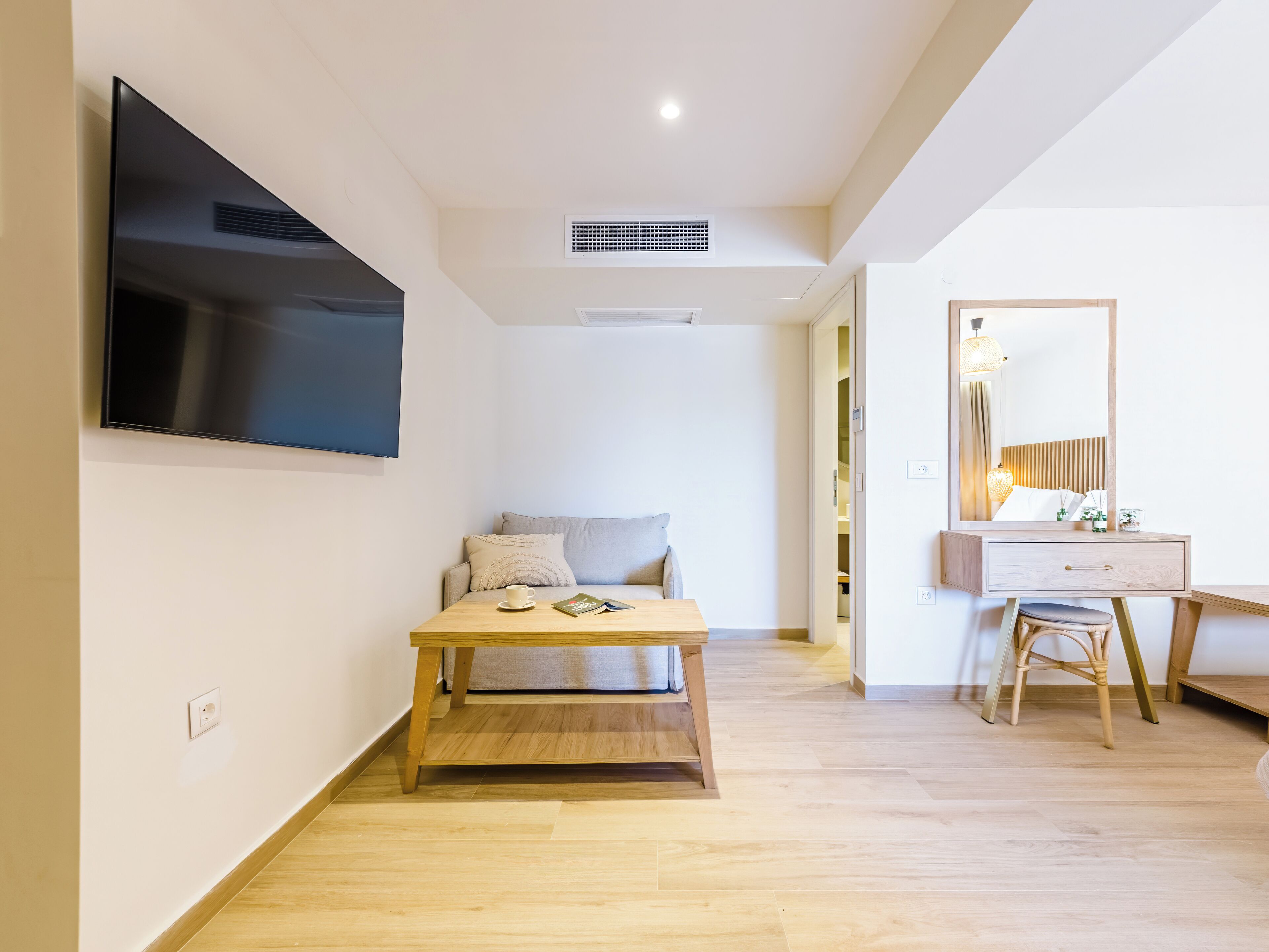 A modern, minimalist bedroom with a flat-screen TV mounted on the wall, a wooden bedside table with a book and a cup, a grey sofa with a pillow, and a wooden dresser with a mirror and lamp.