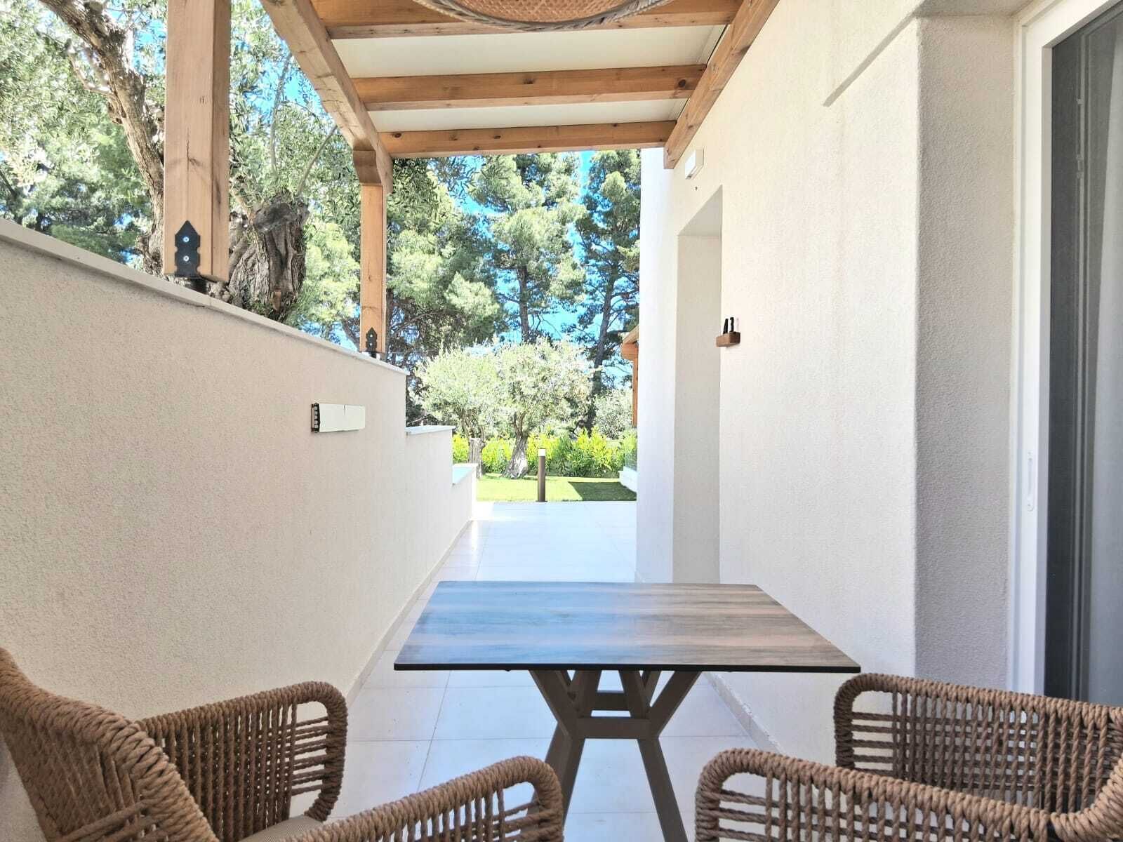 A covered patio with wicker chairs and a table, overlooking a garden with trees and a lawn.
