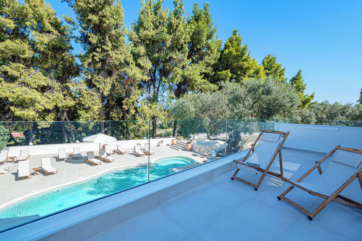 A rooftop pool area with lounge chairs and a glass balustrade overlooking a pool and trees.
