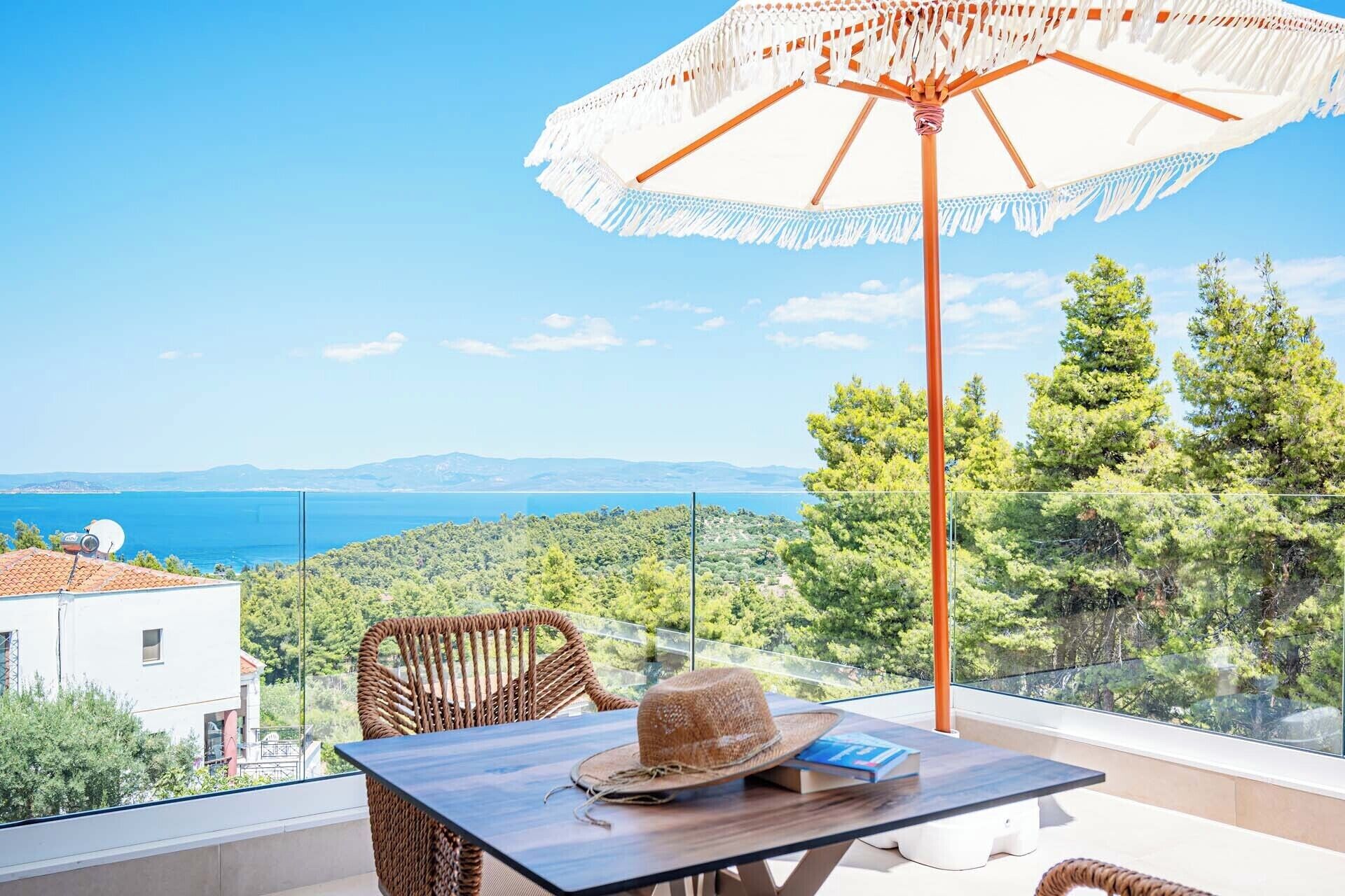 A balcony with a table, chairs, and an umbrella overlooking a coastal landscape with trees and a view of the sea.