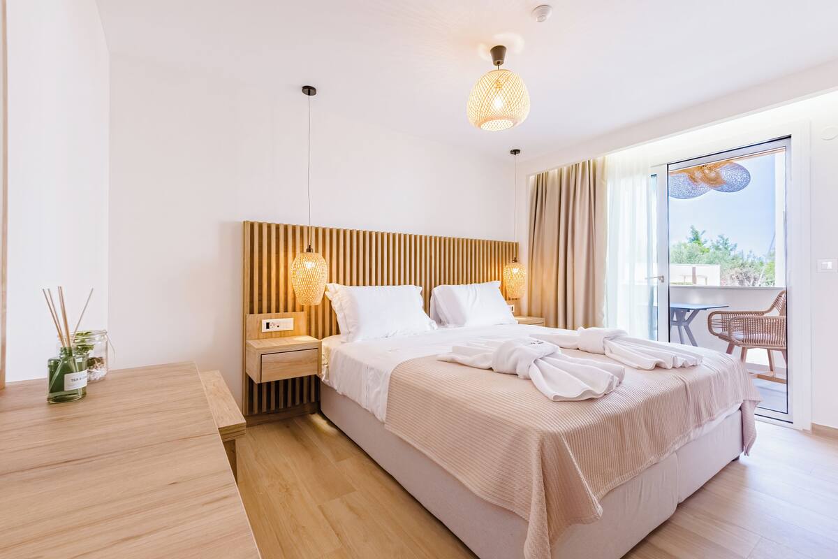 A modern hotel room with a large bed, wooden headboard, bedside tables with lamps, a glass door leading to a balcony, and a small table with a vase and reed diffusers.