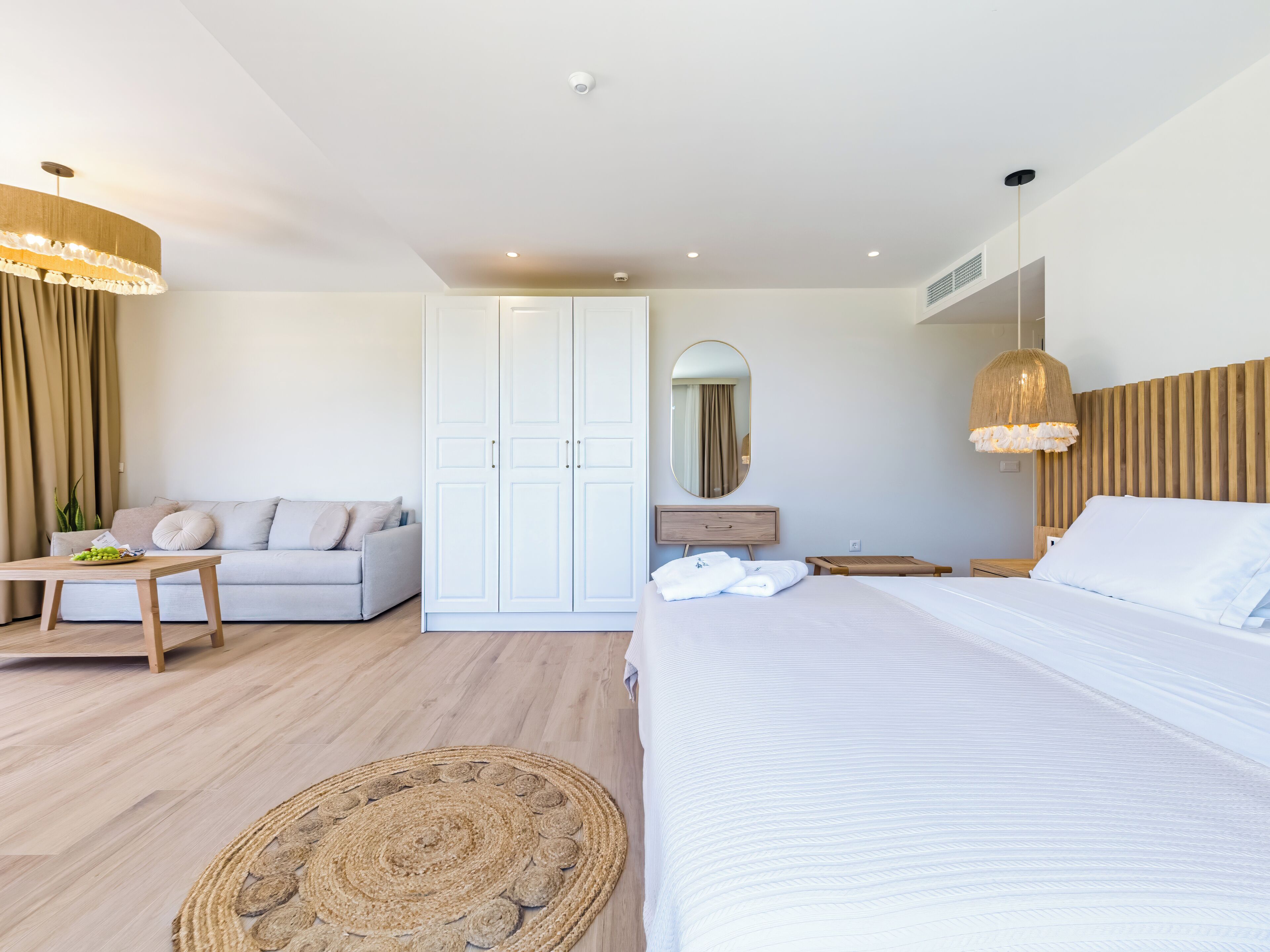 A modern hotel room with a large bed, a sofa, a wooden nightstand, a round woven rug, and a large mirror.