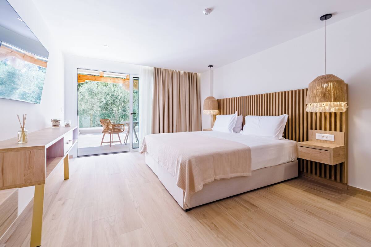 A modern hotel room with a large bed, a desk, and a view of greenery outside.