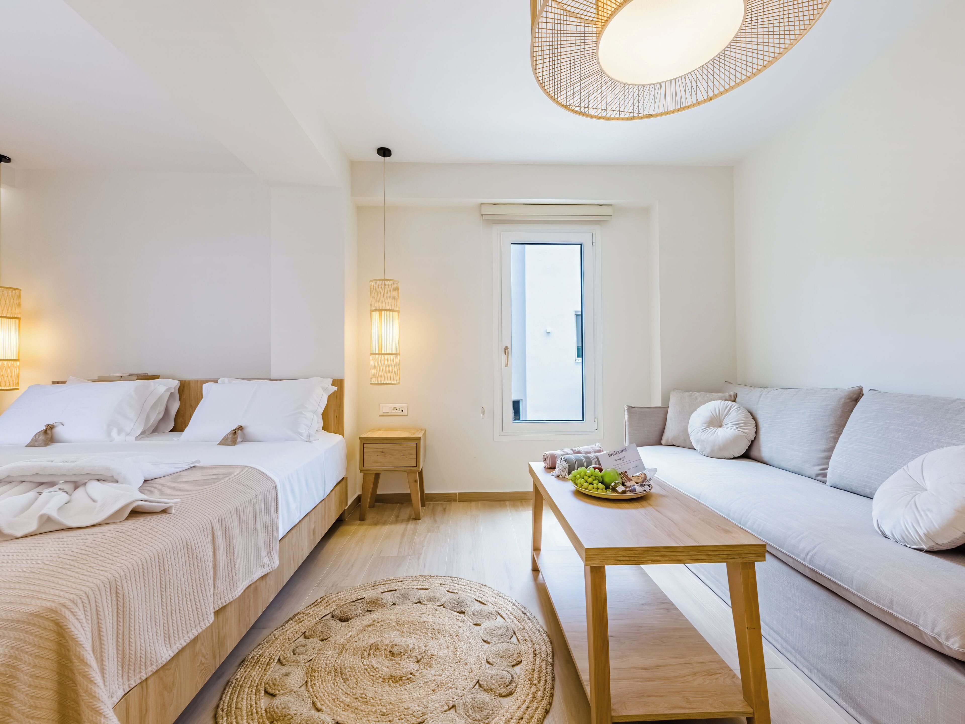 Une chambre d’hôtel moderne, équipée d’un lit, d’un canapé, d’une petite table avec des fruits, d’un tapis tissé et d’une fenêtre donnant sur un paysage agréable.