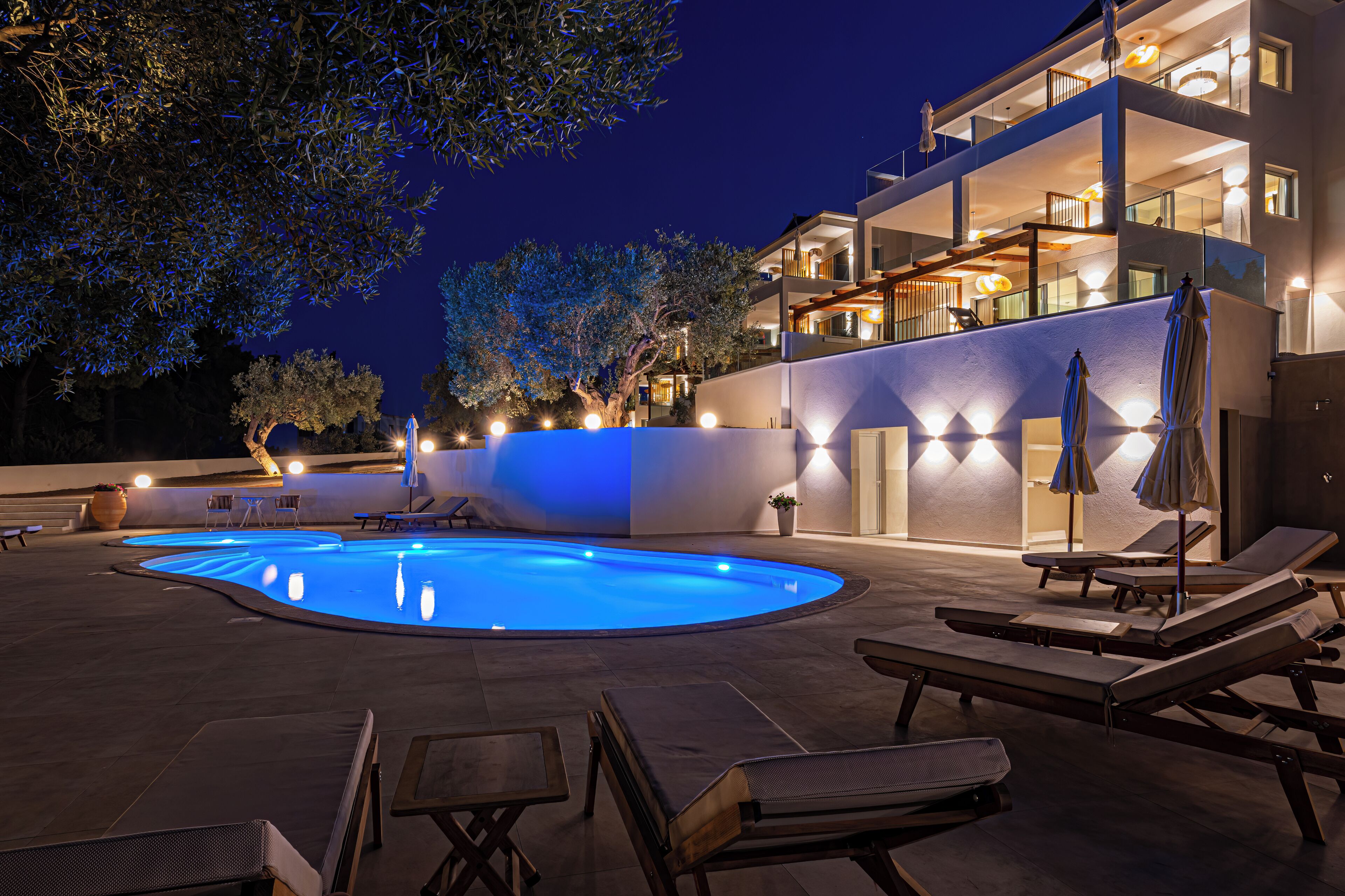 Una piscina con sillas de descanso y mesas, rodeada por un edificio con varios balcones e iluminación exterior.