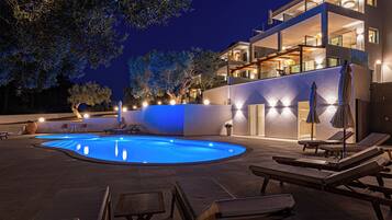 Una piscina con sillas de descanso y mesas, rodeada por un edificio con varios balcones e iluminación exterior.