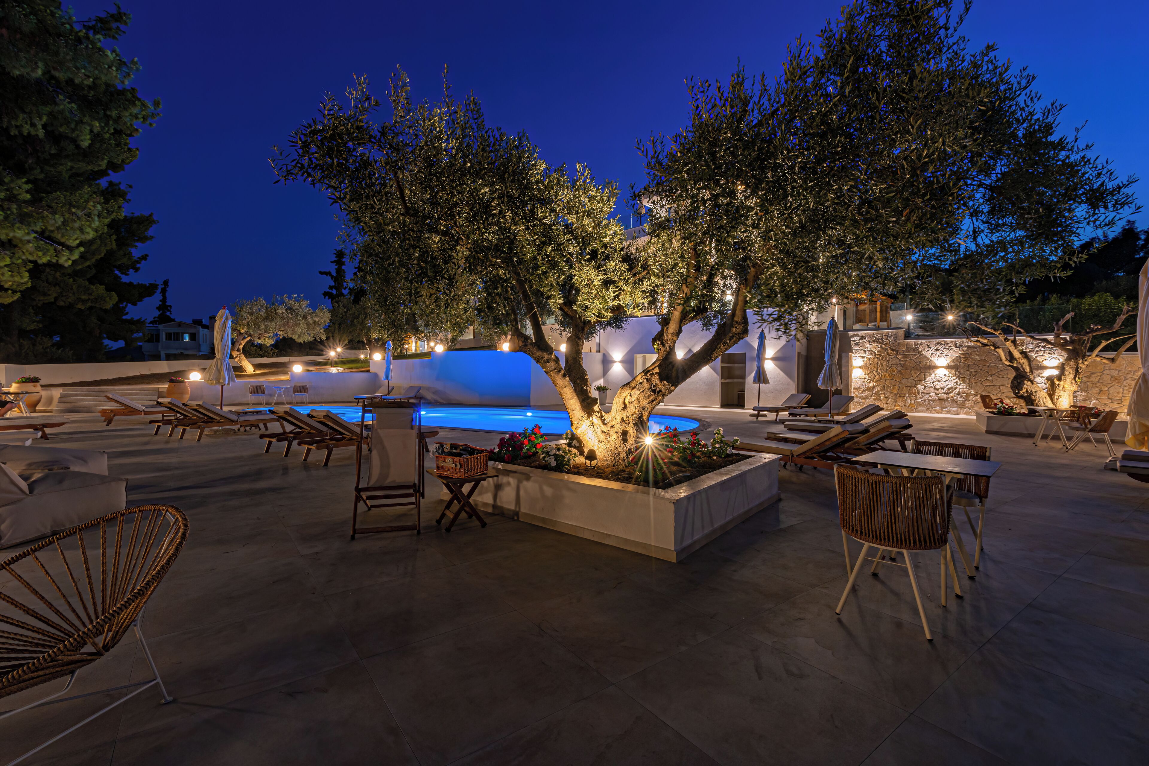 Zona de piscina al aire libre con sillones de descanso, un árbol y un muro de piedra.