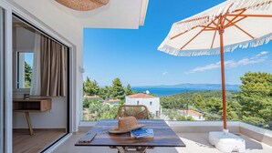 Un balcón con una mesa, una silla de mimbre, un sombrero y una sombrilla sobre una vista panorámica de casas y el mar.
