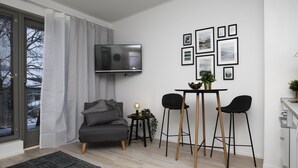 En moderne stue med en grå sofa, et lille bord med to sorte stole, et fjernsyn ophængt på væggen og billeder i rammer over bordet.