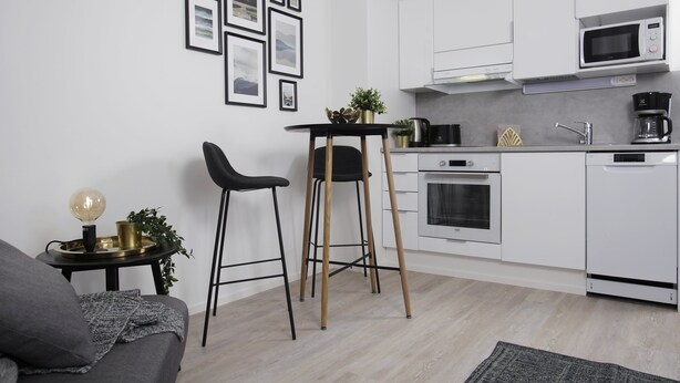 Et moderne køkken med hvide skabe, en mikroovn, ovn og kaffemaskine. Der er to sorte barstole og et lille bord med planter.
