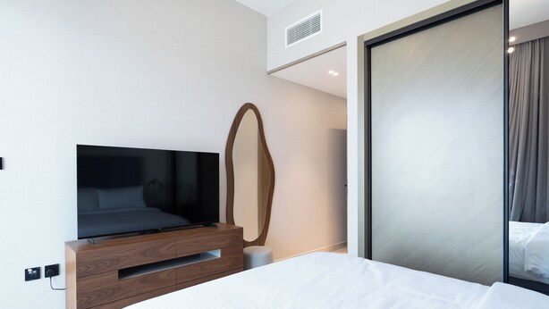 A modern hotel room with a large bed, a wooden dresser, a flat-screen TV, and a large mirror. - Spacious 2BR Canal View - Heart of B Bay (Dubai)