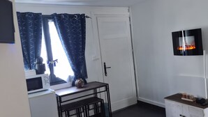 Et lille, godt oplyst rum med en hvid dør, et fjernsyn monteret på væggen, en mikrobølgeovn på en bordplade og et lille bord med en vase.
