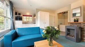 Ein Wohnzimmer mit blauem Sofa, Couchtisch und Kamin. Es gibt eine Küchenzeile mit weißen Schränken und einem Fenster mit Vorhängen.