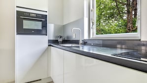 A modern kitchen with white cabinets, a black countertop, a built-in oven, a stainless steel sink, and a window with a view of trees. - Detached 4-person villa (Steendam)