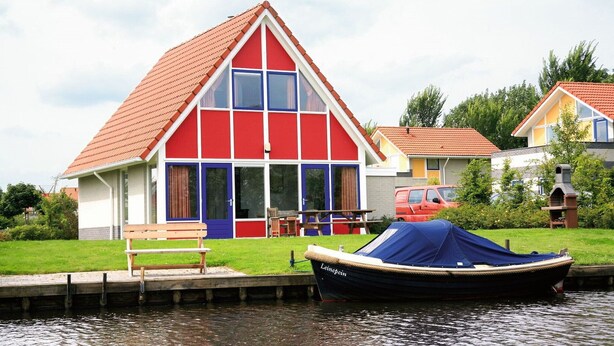 A red and white house with a blue door, a wooden bench, and a boat named "Leinecim" docked by the water. - Holiday villa for 4 persons (Steendam)