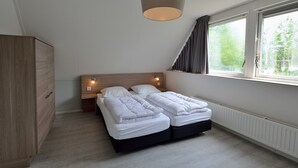A bedroom with a wooden wardrobe, a double bed with white bedding, a headboard with wall-mounted lights, and a window with curtains. - Holiday villa for 4 persons (Steendam)