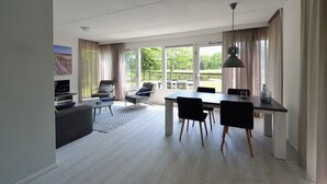A modern living room with a sofa, a coffee table, and a dining area with a table and chairs. There is a TV on a stand and a window with curtains. - Holiday villa for 4 persons (Steendam)