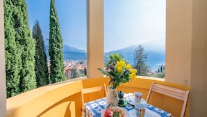 A balcony with a table set for two, a vase with flowers, and a view of a coastal town and mountains.