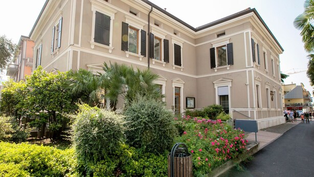 Ein zweistöckiges Gebäude mit einer symmetrischen Fassade, großen Fenstern mit Fensterläden und einem gepflegten Garten mit verschiedenen Sträuchern und blühenden Pflanzen.