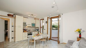 A modern kitchen with white cabinets, a dining table set for four, and a grey sofa in the living area. - Appartamento Lara in Riva del Garda (Riva del Garda)