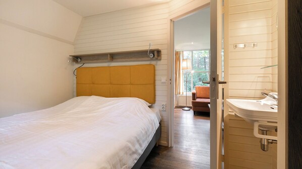 Un dormitorio con cama, cabecero de madera, mesita de noche y baño con lavamanos y espejo.