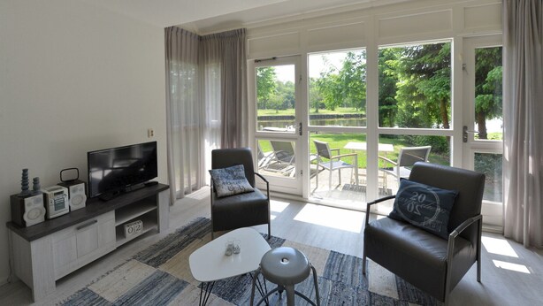 A modern living room with a flat-screen TV, a coffee table, two armchairs, and a sofa. There is a large sliding glass door leading to an outdoor patio with a pool and greenery. - Bungalow for 4 persons (Steendam)