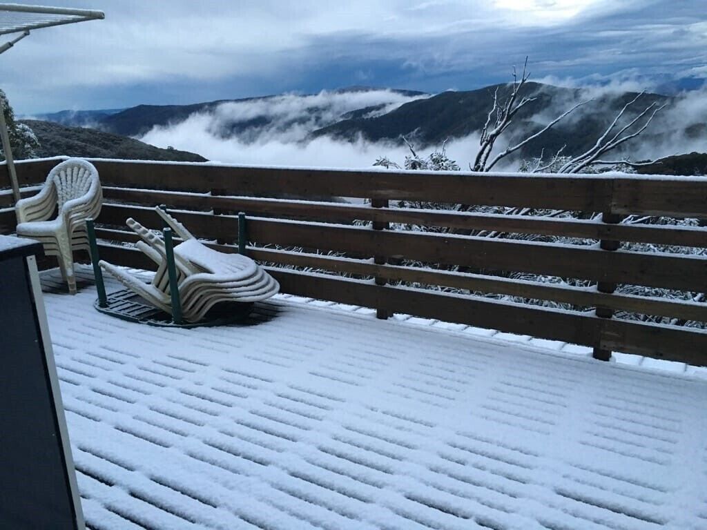 A snow-covered outdoor seating area with a bench and chairs, overlooking a mountainous landscape with clouds.