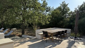 Eine Terrasse im Freien mit einem Holztisch und Stühlen, umgeben von Bäumen und einer überdachten Sitzgelegenheit.