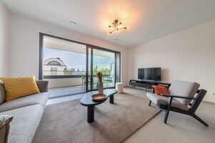 Un salón moderno con sofá, mesa de centro, silla, televisor de pantalla plana y un amplio ventanal con vistas a edificios y árboles.