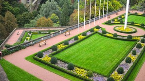 Un jardín formal con césped central, parterres de flores y un puente al fondo.
