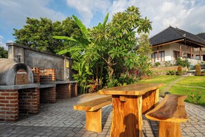 An outdoor barbecue area with a brick oven, wooden benches, and a garden with various plants and a house in the background. - Villa Linggah Longgah, Bali - Spectacular Clifftop Elegance! (Kabupaten Buleleng)