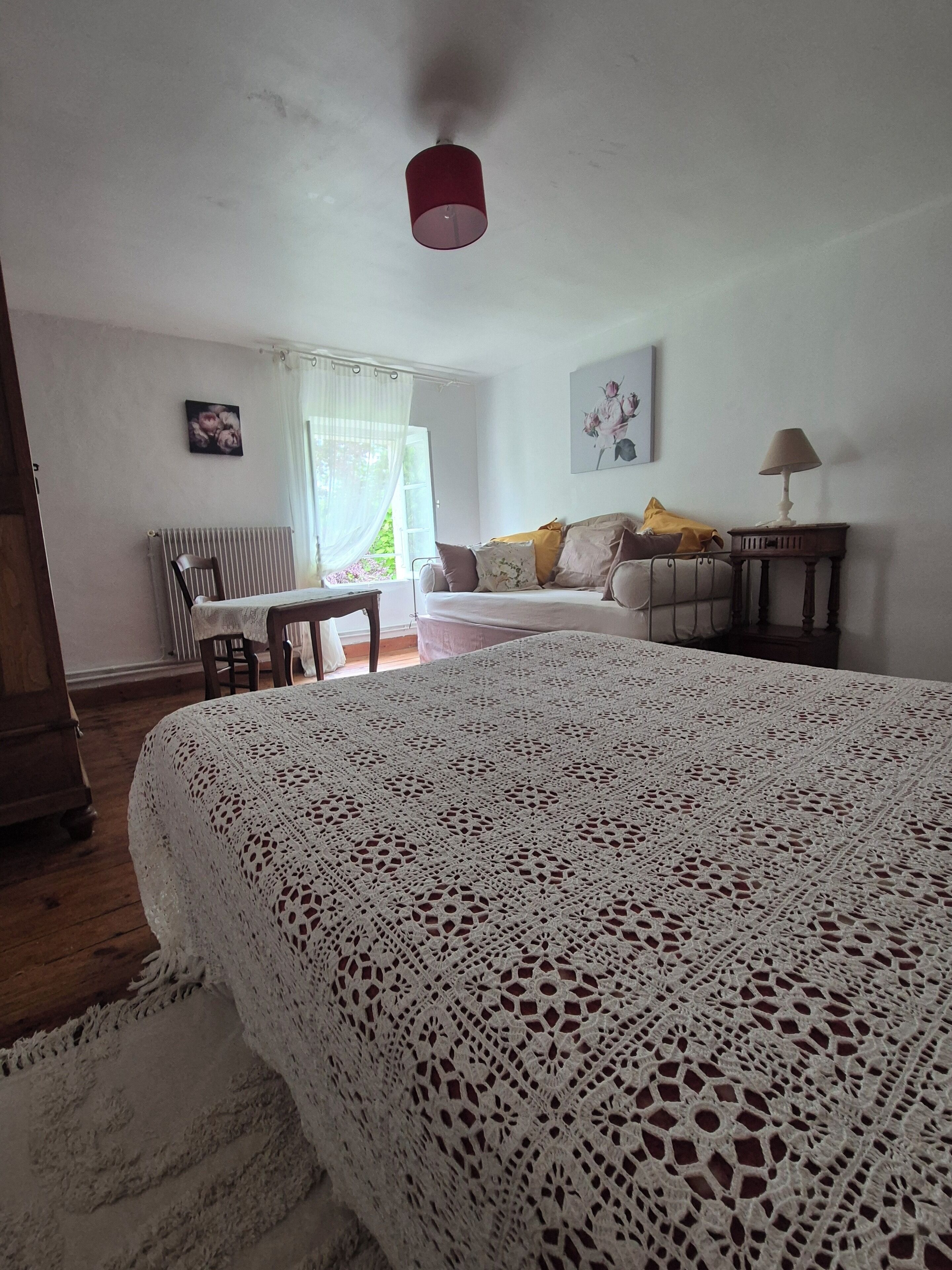 A bedroom with a large bed, a wooden dresser, a table, and a chair. There is a window with curtains, a radiator, and a lamp on a side table.