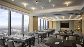 A modern hotel dining area with multiple tables, chairs, and a large TV screen.