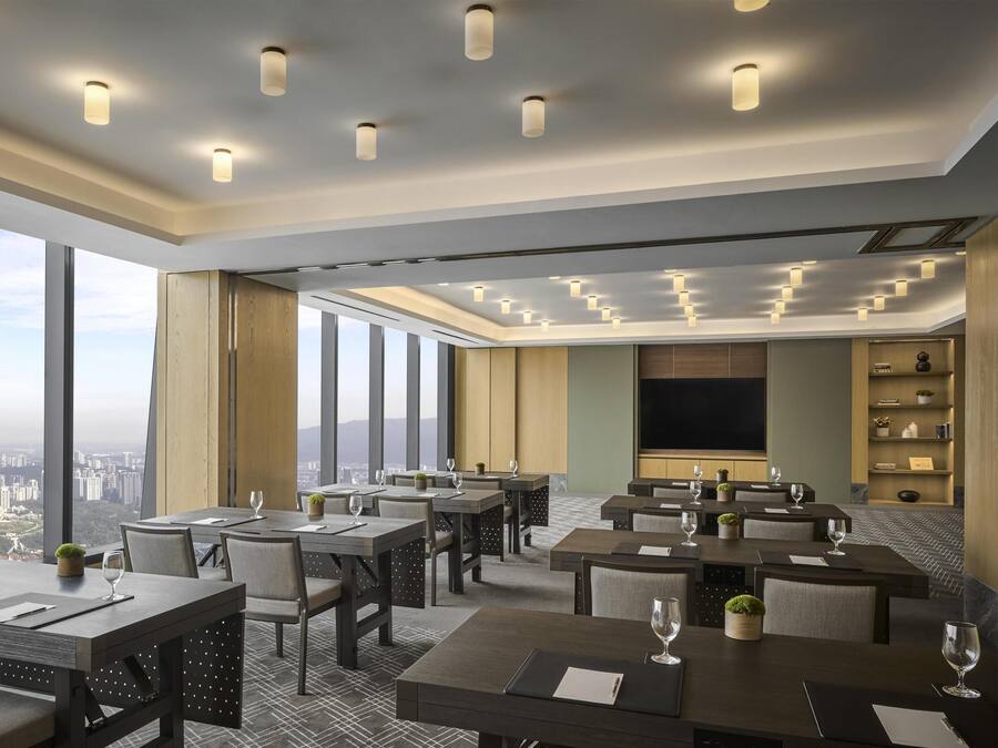 A modern hotel dining area with multiple tables, chairs, and a large TV screen.